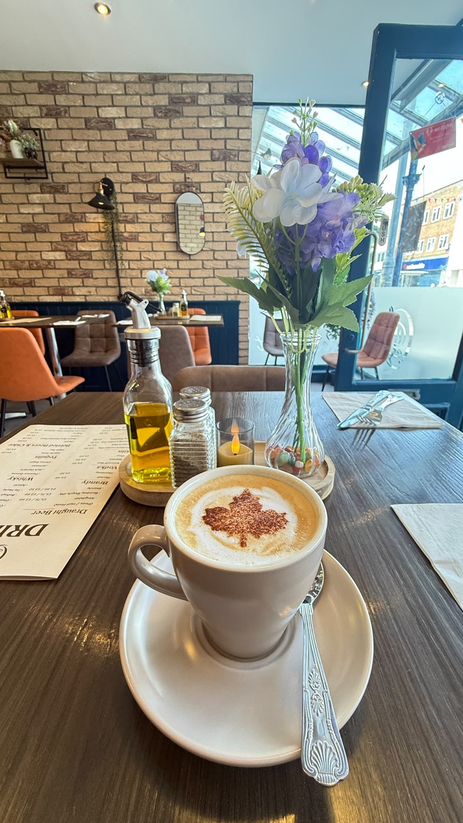 A latte with a maple-leaf design in the foam on a table in the sunlit dining room, with brick walls, orange chairs and fresh flowers